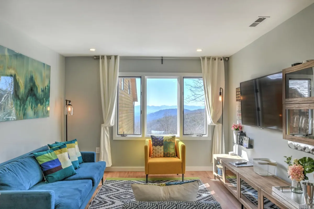 The image showcases a bright, inviting living space with a large window framing breathtaking mountain views. Inside, a plush blue sofa and vibrant yellow accent chair create a cozy, stylish spot to relax or socialize. The warm sunlight streams into the room, highlighting tasteful artwork and inviting guests to unwind while enjoying the natural beauty just beyond the glass.