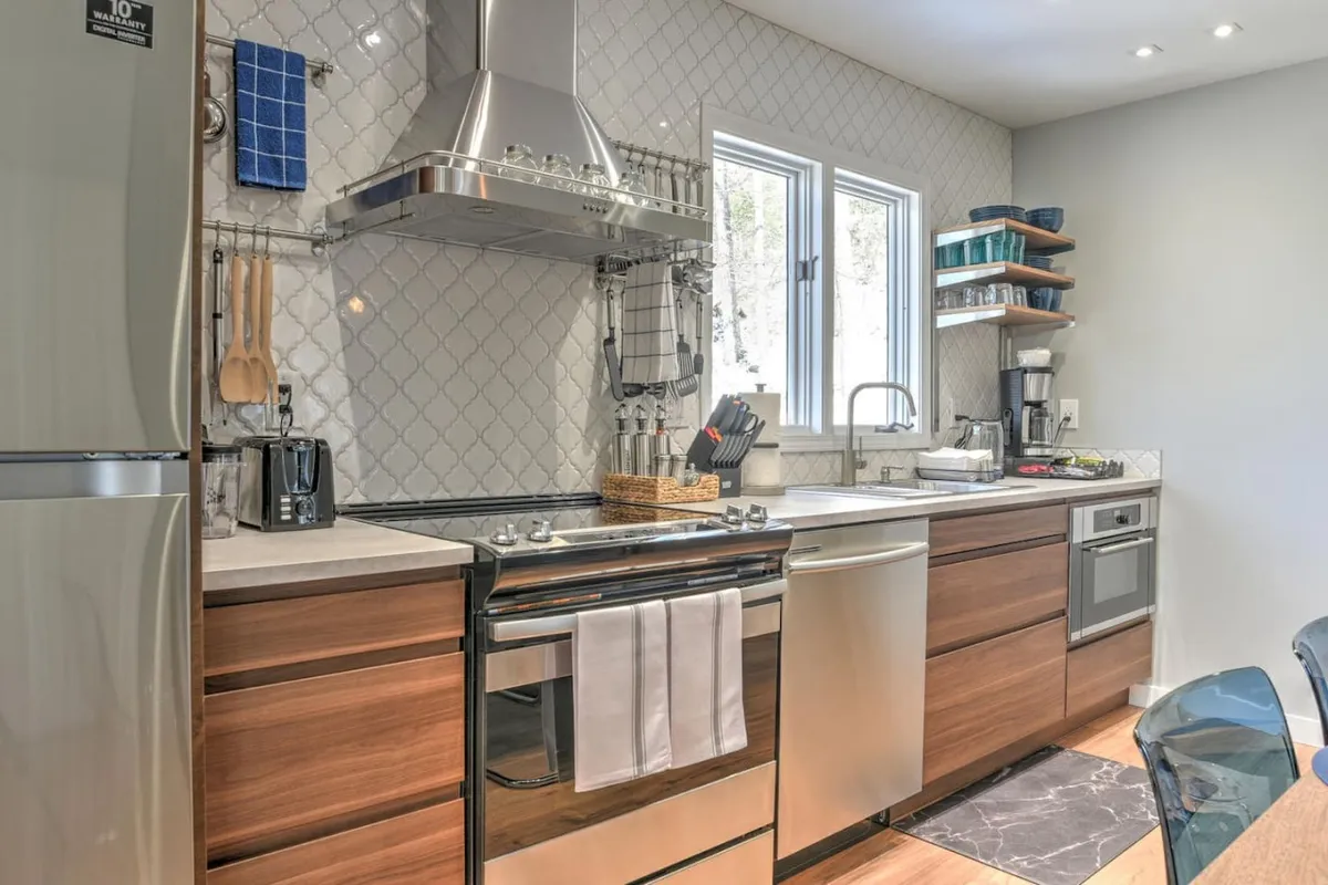 A spacious, modern kitchen featuring stainless steel appliances, wooden cabinetry, a central kitchen island, and open shelving. Natural light streams in through a large window, highlighting the elegant surfaces and contemporary design. The space feels welcoming and is set up for both casual meal prep and entertaining.
