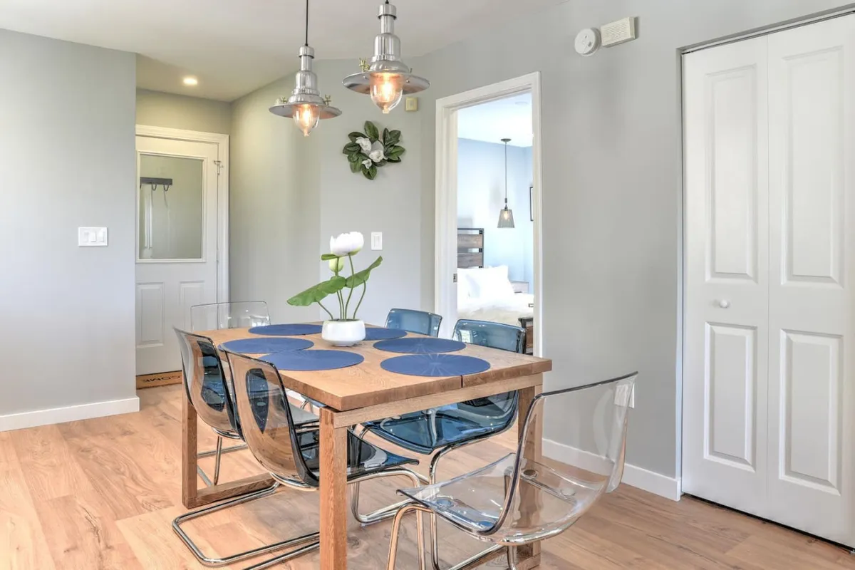 A modern dining area with a contemporary table set for a meal, accented by blue placemats and decorative plants. Stylish hanging light fixtures provide a relaxed ambiance, ideal for intimate dinners or celebratory toasts.