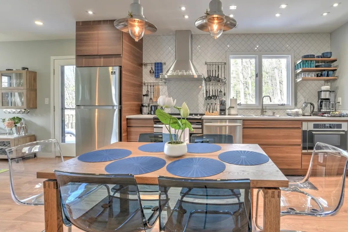 A stylish dining area with a modern table set with blue placemats, adjacent to the kitchen. Large windows flood the room with natural light, offering scenic views. Contemporary hanging light fixtures add a warm, inviting touch, and the kitchen’s state-of-the-art appliances are visible in the background, seamlessly connecting both spaces.