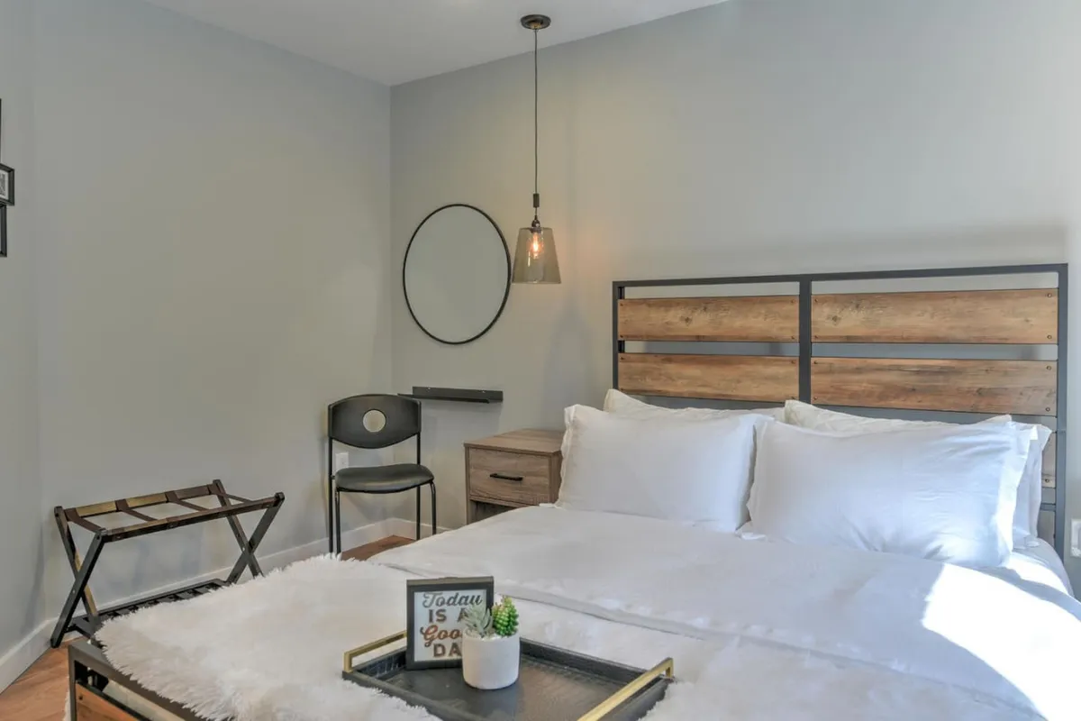 A serene bedroom suite featuring a plush bed with crisp white linens, a wooden headboard, a modern light fixture overhead, a circular mirror on the wall, and a small desk with a chair. The space is flooded with gentle natural light, creating a peaceful and cozy atmosphere.