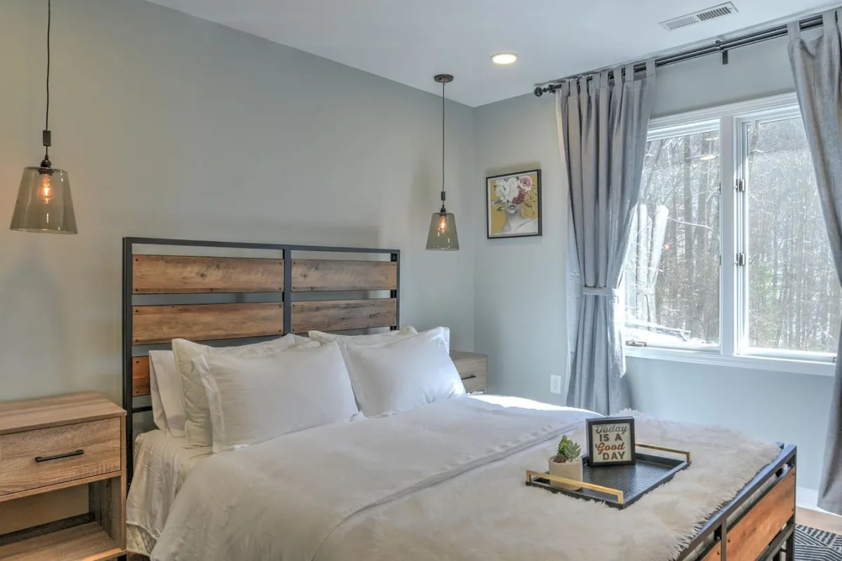 A tranquil bedroom with a plush bed dressed in soft white bedding, a wooden headboard, a nightstand with decorative items, framed artwork above the bed, and curtains gently allowing in natural light. The setting feels restful and inviting, ideal for unwinding.