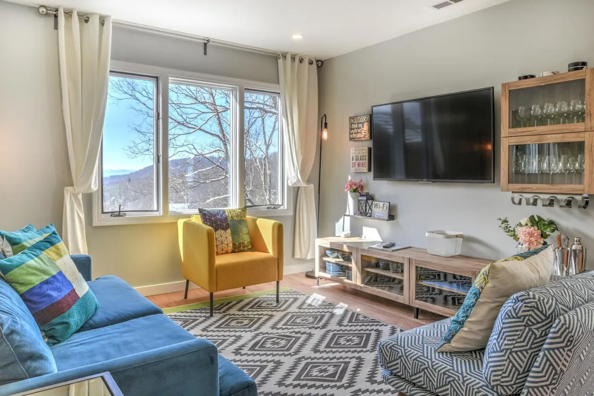 A spacious living area featuring large windows that reveal striking mountain views, colorful accent chairs, a textured area rug, and a contemporary media console. The room is flooded with natural light, creating a bright and inviting atmosphere ideal for relaxation and social gatherings.