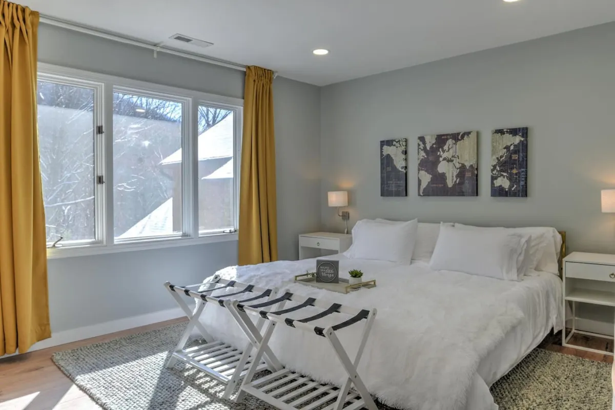A serene bedroom featuring a plush white bed, soft yellow curtains framing the window, warm nightstand lamps, and a world map artwork above the headboard. Natural morning light fills the space, creating a cozy and inviting retreat.